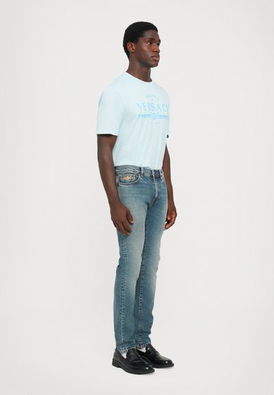 Light blue cotton t-shirt with "Versace" text, paired with faded denim jeans and black leather shoes. Model stands side profile against a white background.