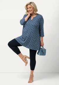 Blå tunika med blommigt mönster, trekvartsärmar, kombinerad med svarta leggings och beige klacksandaler med remmar. Håller en ljusblå handväska.