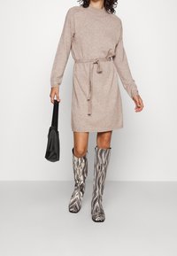 Abito a maglia beige con collo rotondo e cintura in vita, abbinato a stivali al ginocchio a motivo in bianco e nero e una borsa a spalla nera.