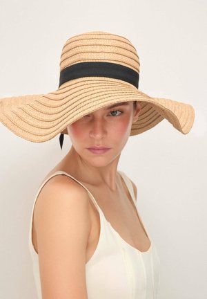 Frau mit heller Haut und blauen Augen, die einen breitkrempigen beigen Strohhut mit schwarzem Band und ein weißes ärmelloses Oberteil trägt und seitlich blickt.