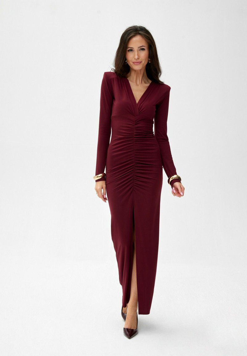 Burgundské šaty s dlouhým rukávem a řasením, s hlubokým výstřihem do V a rozparkem až po stehno. Hladká tkanina, přiléhavý střih, doplněné špičatými podpatky.