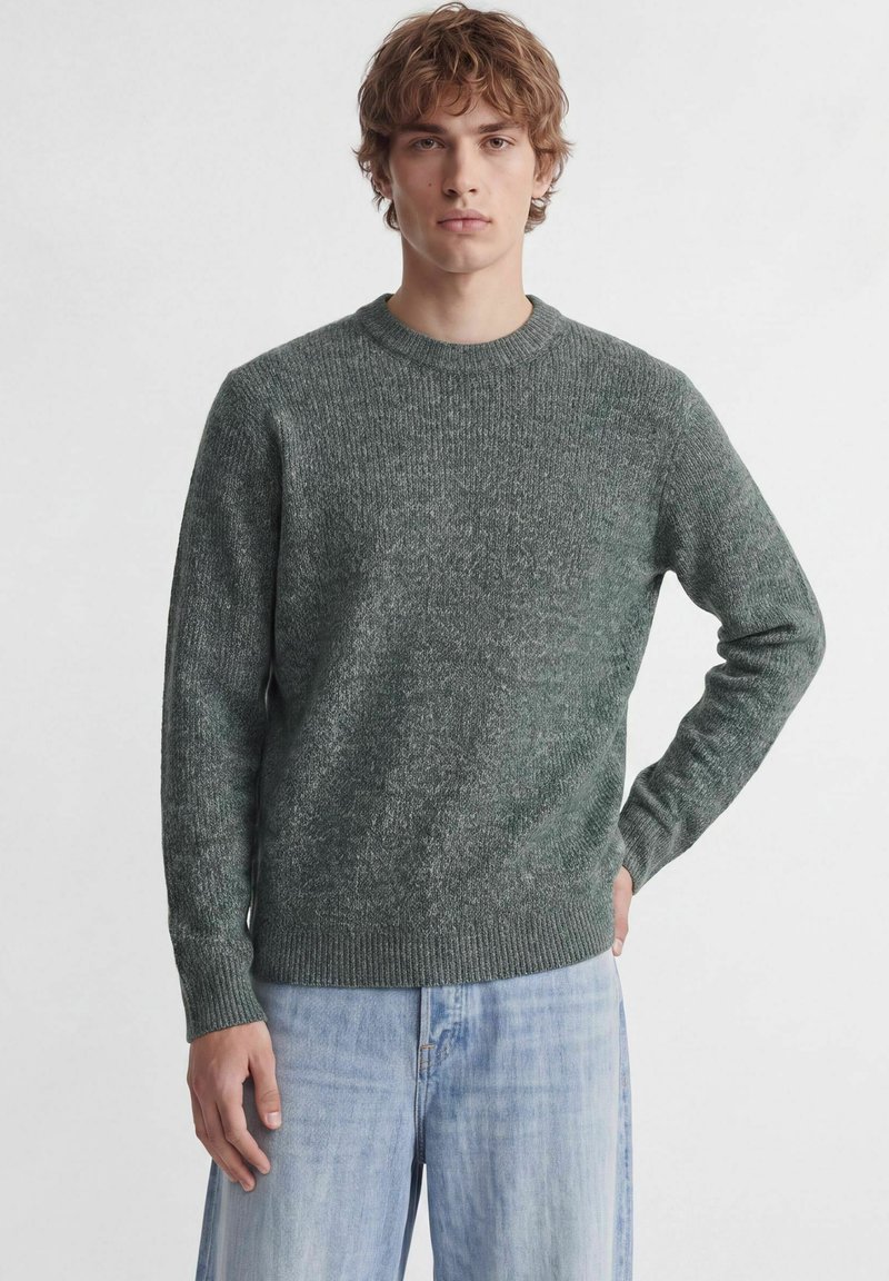 Jeune homme aux cheveux châtain clair bouclés, portant un pull en maille gris foncé et un jean bleu clair, debout avec une main sur la hanche.