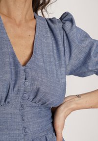 Niebieska sukienka o teksturowanej powierzchni, z głębokim dekoltem w kształcie litery V, bufkowymi rękawami i sześcioma guzikami z przodu. Dopasowana talia i luźna spódnica.