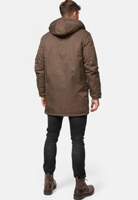 Bruine, capuchonparka met een gladde textuur, voorzien van een langere snit en versterkte stiksels. Gecombineerd met donkere jeans en bruine laarzen.