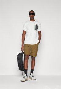 Hvit t-skjorte med en grønn lomme, beige cargobukser, svart ryggsekk og lyse joggesko. Tilbehør inkluderer mørke solbriller og svarte sokker.