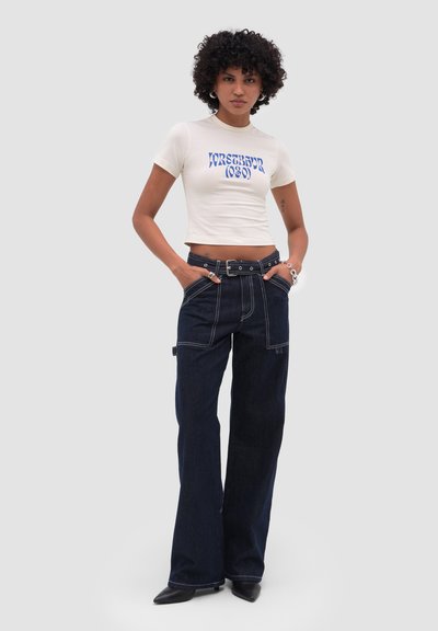 Femme aux cheveux bouclés portant un t-shirt court blanc avec un texte bleu et un jean large foncé avec une ceinture, debout les mains dans les poches.