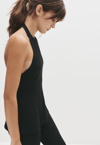 Sort halterneck-top med en løs pasform, lavet af et blødt stof. Har en buet kant og uden ærmer, parret med sorte leggings.