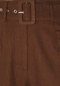 Pantalon en lin marron avec une large ceinture à boucle carrée, coutures visibles et tissu texturé. Design classique avec plis à l'avant.