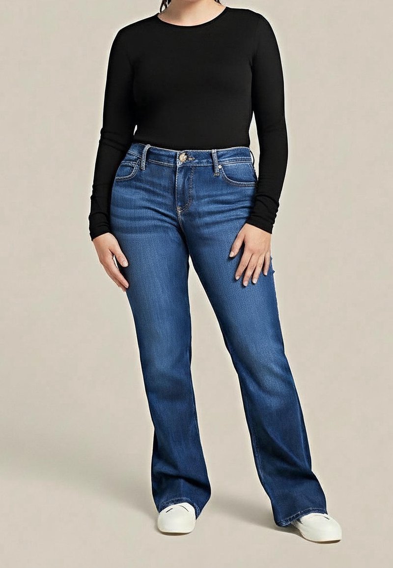 Femme portant un haut noir cintré à manches longues, un jean bootcut bleu et des baskets blanches, debout devant un fond uni.
