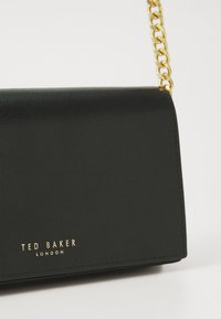 Bolsa de couro preto com superfície texturizada, alça de corrente dourada e logotipo em relevo "TED BAKER LONDON" em dourado na frente.