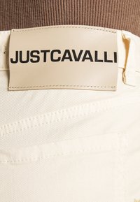 Pantalones de mezclilla blanca con una etiqueta de cuero que lleva "JUSTCAVALLI" en letras negras en negrita, destacando los detalles de las costuras y los bolsillos.