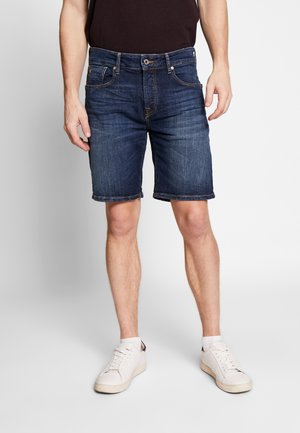 Man wearing dark blue denim shorts, white sneakers, and a dark top, standing against a plain light background.
