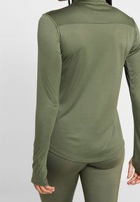 Olivegrünes Langarmoberteil mit hohem Kragen, glatter Textur, taillierter Passform und Daumenlöchern an den Ärmeln, kombiniert mit passenden Leggings.