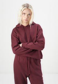 Maroon kročeni dukserica s kapuljačom na vezanje i rebrastim manžetama, uparena s odgovarajućim donjim dijelom hlača na vezanje. Mekana, teksturirana tkanina.