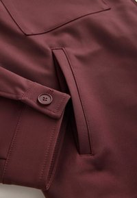 Burgundy jacket with smooth fabric, featuring a buttoned cuff, a patch pocket, and precise stitching along the edges.