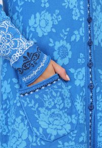 Blauer Strickcardigan mit floralen Mustern, strukturierten Ärmeln mit Akzenten und einer Fronttasche. Abgeschlossen mit blauen Knöpfen entlang der Knopfleiste.