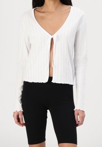 Vit ribbad cardigan med en djup V-ringning, framknäppning med knytning och vågig nederkant. Långa ärmar med texturerad detalj. Bärs över svarta shorts.