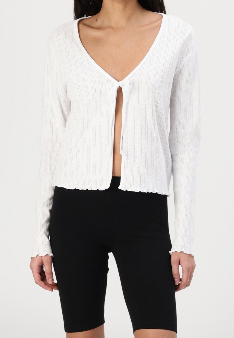 Vit ribbad cardigan med en djup V-ringning, framknäppning med knytning och vågig nederkant. Långa ärmar med texturerad detalj. Bärs över svarta shorts.