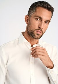 Camicia bianca con colletto strutturato, maniche lunghe e polsini con bottone. Tessuto dalla texture liscia. Il modello aggiusta il colletto con una mano.