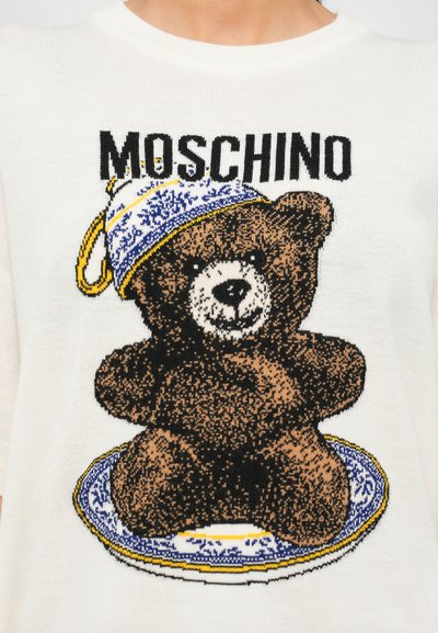 Maglione bianco lavorato a maglia con una grafica di un orso che tiene un piatto decorato blu e oro; l'orso indossa un cappello decorativo nei colori nero e marrone.
