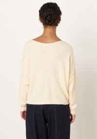 Kvinna med lockigt hår i en låg knut, iklädd en krämfärgad tröja och mörka byxor med smala ränder, vänd bort från kameran mot en enfärgad bakgrund.