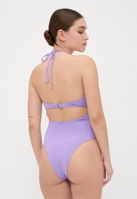 Maillot de bain une pièce violet avec un col halter et une découpe au dos, présentant une fermeture à nouer et un branding subtil près de la taille. Texture lisse.