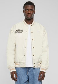 Creme Bomberjacke mit Rippbündchen und Kragen, ausgestattet mit Druckknöpfen und gesticktem Logo auf der linken Brust. Trägt man über einem weißen T-Shirt.