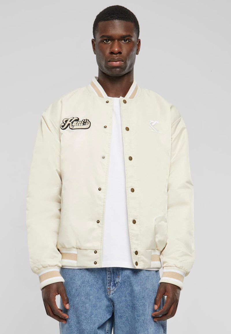 Creme Bomberjacke mit Rippbündchen und Kragen, ausgestattet mit Druckknöpfen und gesticktem Logo auf der linken Brust. Trägt man über einem weißen T-Shirt.