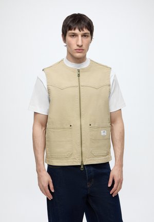 Mand iført en beige ærmeløs lynlåsvest over en hvid kortærmet skjorte og mørkeblå jeans, stående mod en hvid baggrund.