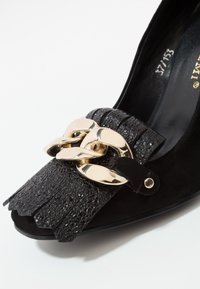 Chaussure pointue texturée noire avec devant frangé et large chaîne dorée brillante sur le dessus.