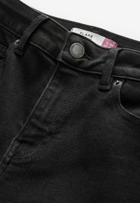 Jeans acampanados negros con una tela texturada, cierre con botón metálico y etiqueta de marca. Presentan un diseño clásico de cinco bolsillos y costuras sutiles.