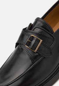 Musta nahast loafers, millel on lai rihm ja metallist pandla aksent. Sile tekstuur kontrastsete õmblustega ja poleeritud viimistlusega.