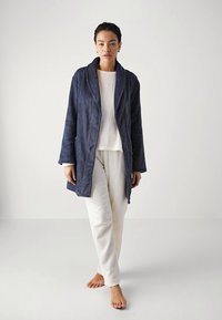Mörkblå fleece-kappa med en jämn textur, sjalkrage, sidofickor och långa ärmar, bärs över en vit topp och ljusa byxor.