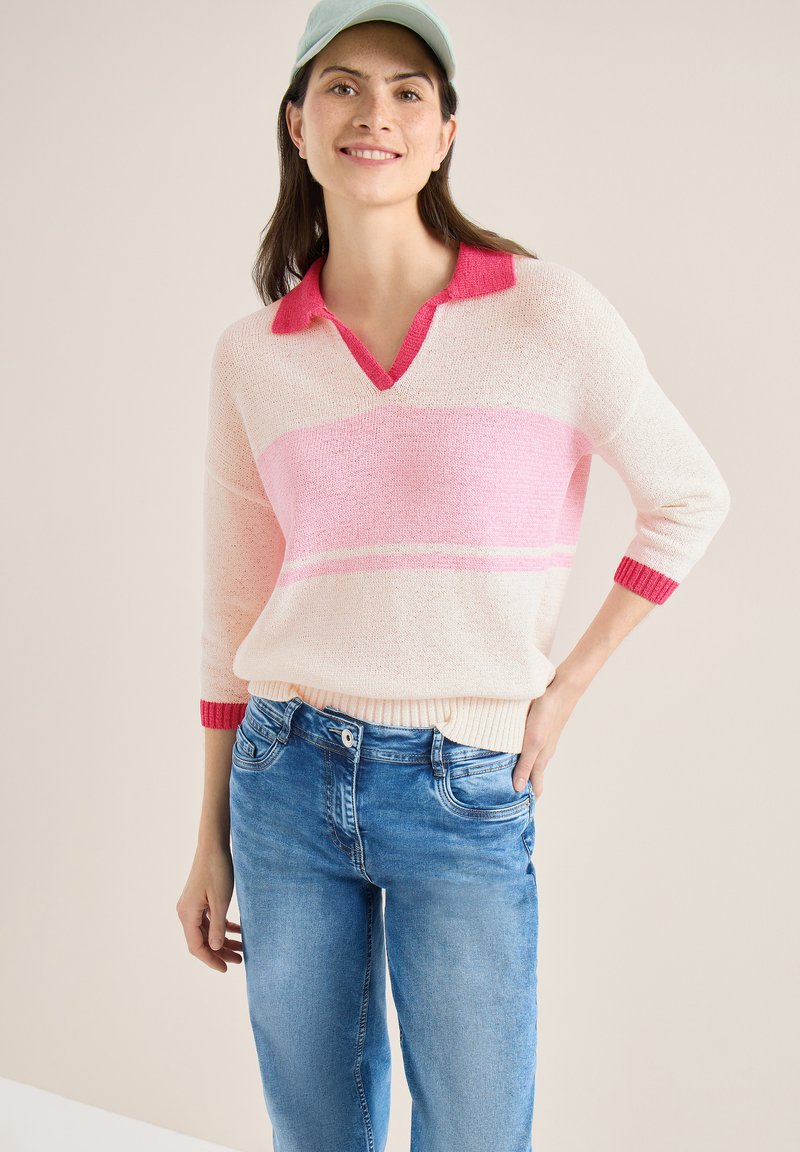 Femme portant un pull à rayures roses et blanches avec un col, un jean bleu et une casquette de baseball vert clair, souriante avec une main sur la hanche.