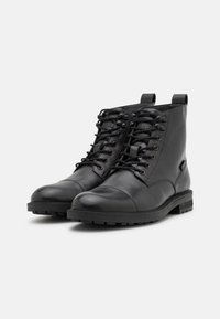 Botas de cuero negro hasta el tobillo con puntas redondas, cordones en la parte frontal, ojetes de metal y suelas de goma resistentes. Presenta dos tiradores de bucle en la parte superior.