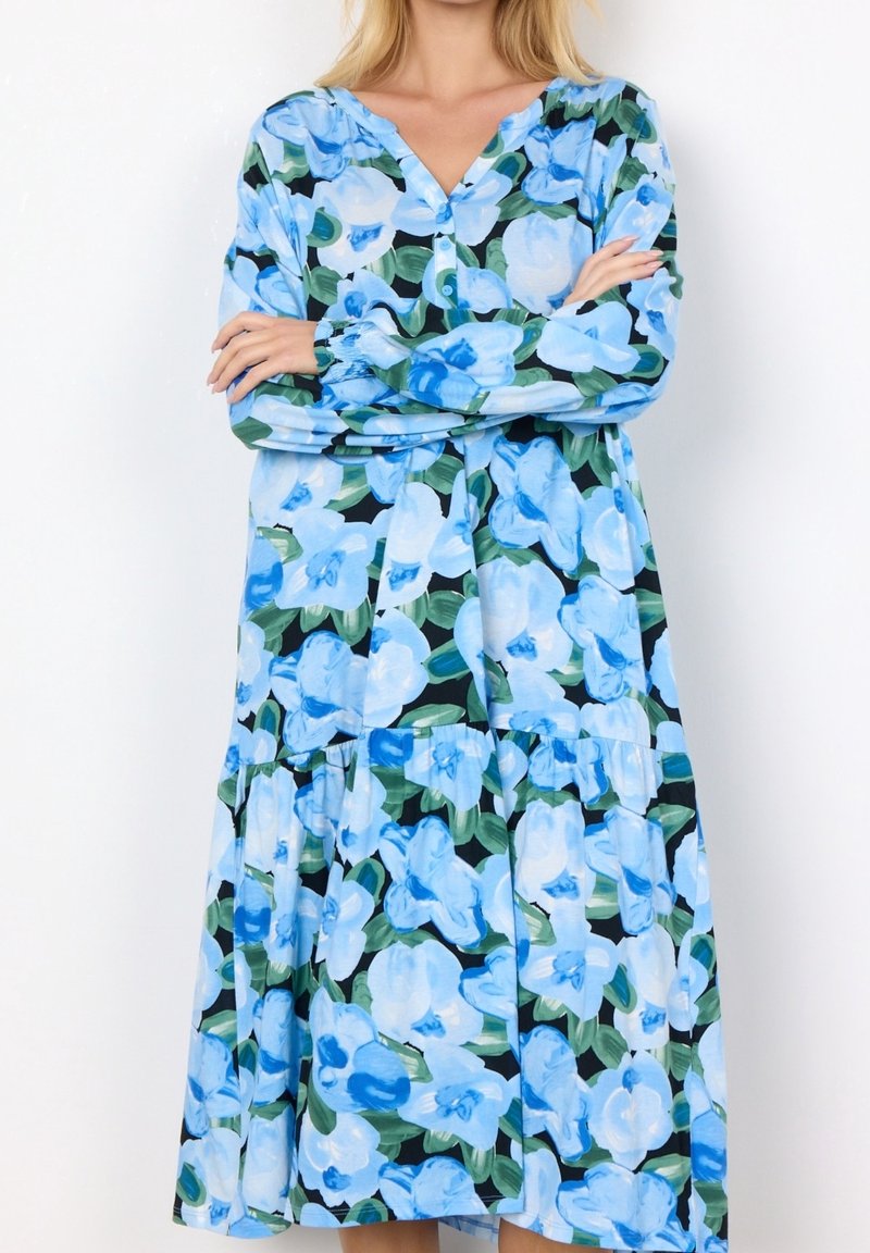 Femme portant une robe à manches longues avec un grand imprimé floral bleu et blanc et des feuilles vertes sur un fond noir, bras croisés.