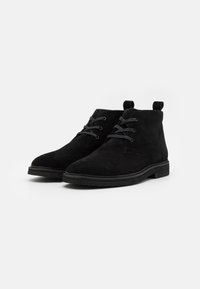 Botines de ante negros con punta redondeada, cordones en tono y tiradores. Presentan una suela de goma plana y un diseño minimalista.