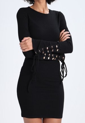 Frau trägt ein eng anliegendes schwarzes Kleid mit langen Ärmeln mit Schnürdetail und Metallösen an den Unterarmen, stehend mit verschränkten Armen.