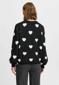 Schwarzer Pullover mit weißen Herzmustern, runder Halsauschnitt und Puffärmeln. Weicher Stoff. Getragen mit grauen Hosen, Rückansicht gezeigt.
