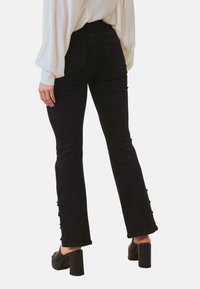 Jeans negros de campana con cintura alta, que cuentan con un detalle de botón en las piernas y un acabado suave y texturizado. Combinados con tacones de plataforma.