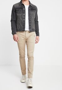 Chaqueta de mezclilla oscura con dos bolsillos en el pecho, camiseta blanca, pantalones beige ajustados y zapatillas blancas. Diseño simple, sin patrones visibles.