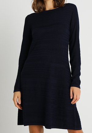 Frau trägt ein langärmliges, knielanges, dunkel marineblaues Strickkleid mit Boot-Ausschnitt vor einfarbigem Hintergrund.