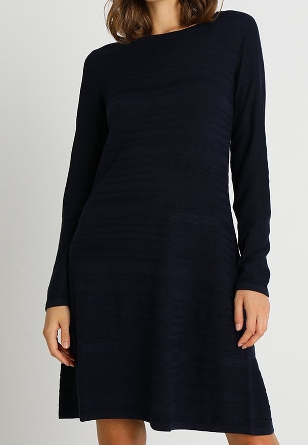 Femme portant une robe en maille bleu marine foncé, manches longues, longueur genou, avec un col bateau, sur un fond uni.