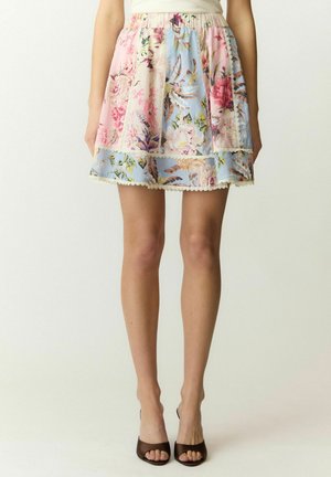 Vrouw die een korte pastelbloemen patchwork rok draagt met kanten bies en bruine open teen hakken, staand tegen een eenvoudige achtergrond.