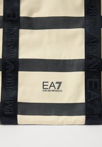 Šviesiai ruda kanvos kuprinė su mėlynomis horizontaliomis juostelėmis, turinti logotipo etiketes ir prekės ženklą, akcentuota tamsiomis tekstūruotomis diržais.