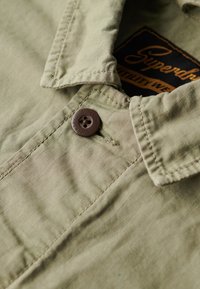 Olive green shirt with a button closure, textured fabric, and a small label displaying the brand name "Superdry" inside the collar.