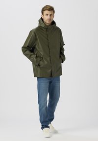 Zöld vízálló dzseki magas gallérral, patent gombokkal és kényelmes szabással, kék farmernadrággal és fehér tornacipővel párosítva. Sima textúra.