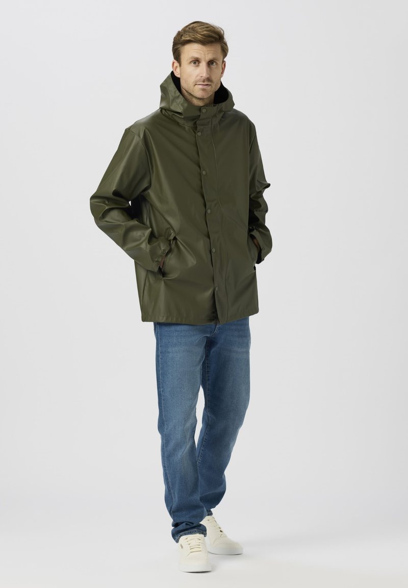 Zöld vízálló dzseki magas gallérral, patent gombokkal és kényelmes szabással, kék farmernadrággal és fehér tornacipővel párosítva. Sima textúra.