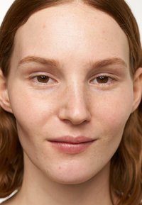 Gros plan sur un visage avec une peau claire, de légères taches de rousseur et des yeux bruns. Texture de peau lisse et cheveux bruns raides. Expression neutre.