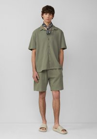 Licht olijfgroene korte mouwen shirt en bijpassende korte broek van gestructureerde stof, gecombineerd met beige slides en een zwart gebloemd sjaaltje om de nek.